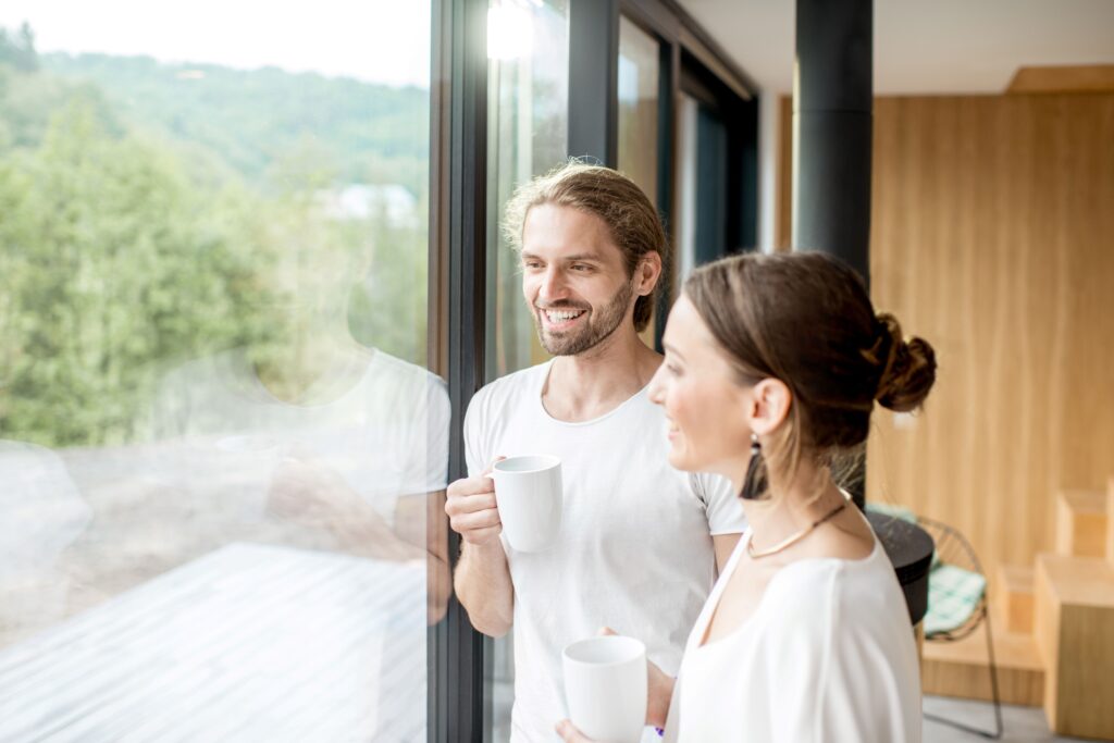 Ein lächelndes Paar steht in einem modernen Zuhause und schaut durch große, klare Fenster nach draußen. Beide halten Tassen in den Händen und genießen die Aussicht.