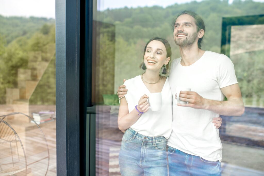 Ein lächelndes Paar steht in einem modernen Zuhause und schaut durch große, klare Fenster nach draußen. Beide halten Tassen in den Händen und genießen die Aussicht.